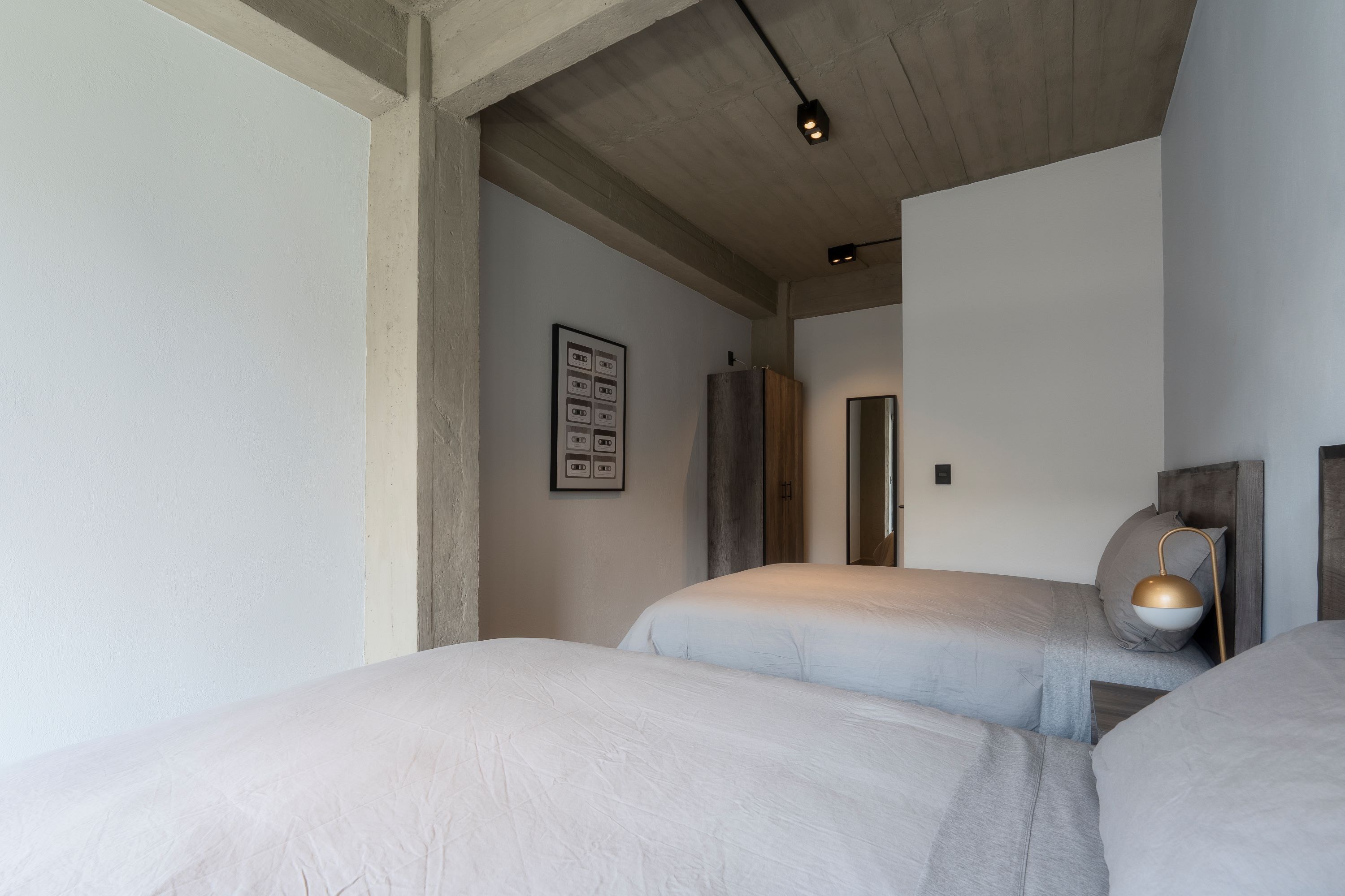 Quarto de hotel moderno com duas camas, cabeceira de madeira, luminária fixada na parede e um quadro emoldurado.