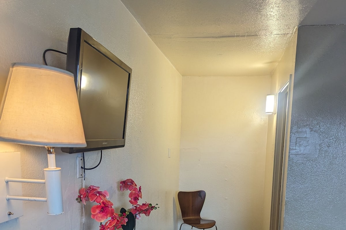A hotel room with a bed, a desk with a lamp, a chair, and a TV mounted on the wall.