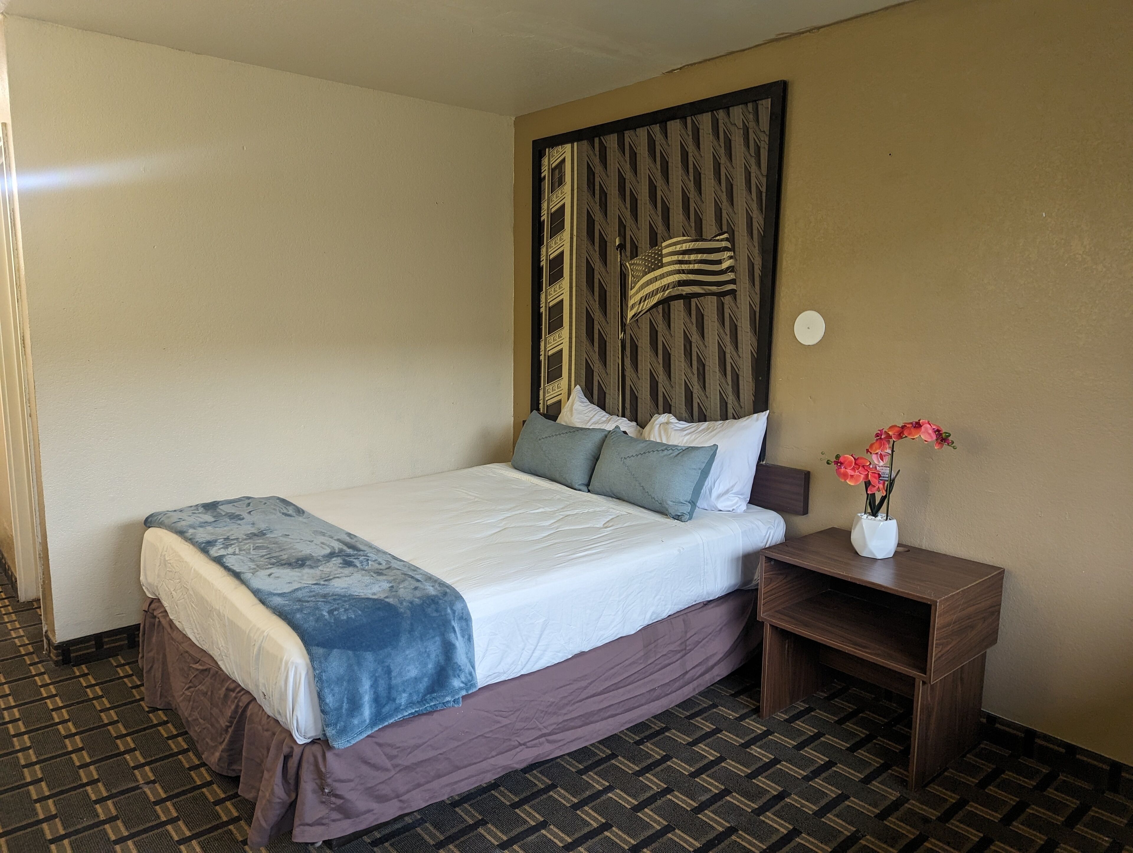 A hotel room with a bed, a nightstand with a vase of flowers, and a wall with a decorative panel.