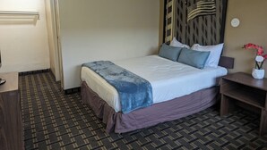 A hotel room with a bed, a nightstand, a desk, and a patterned floor. - Southern Comfort Hotel (Tulsa)