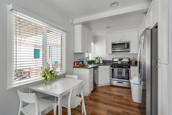 A modern kitchen with white cabinets, a stainless steel oven, a microwave, and a refrigerator. There is a dining table with chairs and a window with blinds. - Entire Complex | Steps To The Sand | Sleeps 20 (Huntington Beach)
