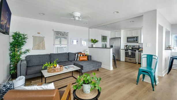 A modern living room with a grey sofa, a coffee table, and a dining area with a teal chair. - Entire Complex | Steps To The Sand | Sleeps 20 (Huntington Beach)