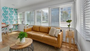 A spacious living room with a leather sofa, a round coffee table, and a dining area with a white chair and a small table. - Entire Complex | Steps To The Sand | Sleeps 20 (Huntington Beach)