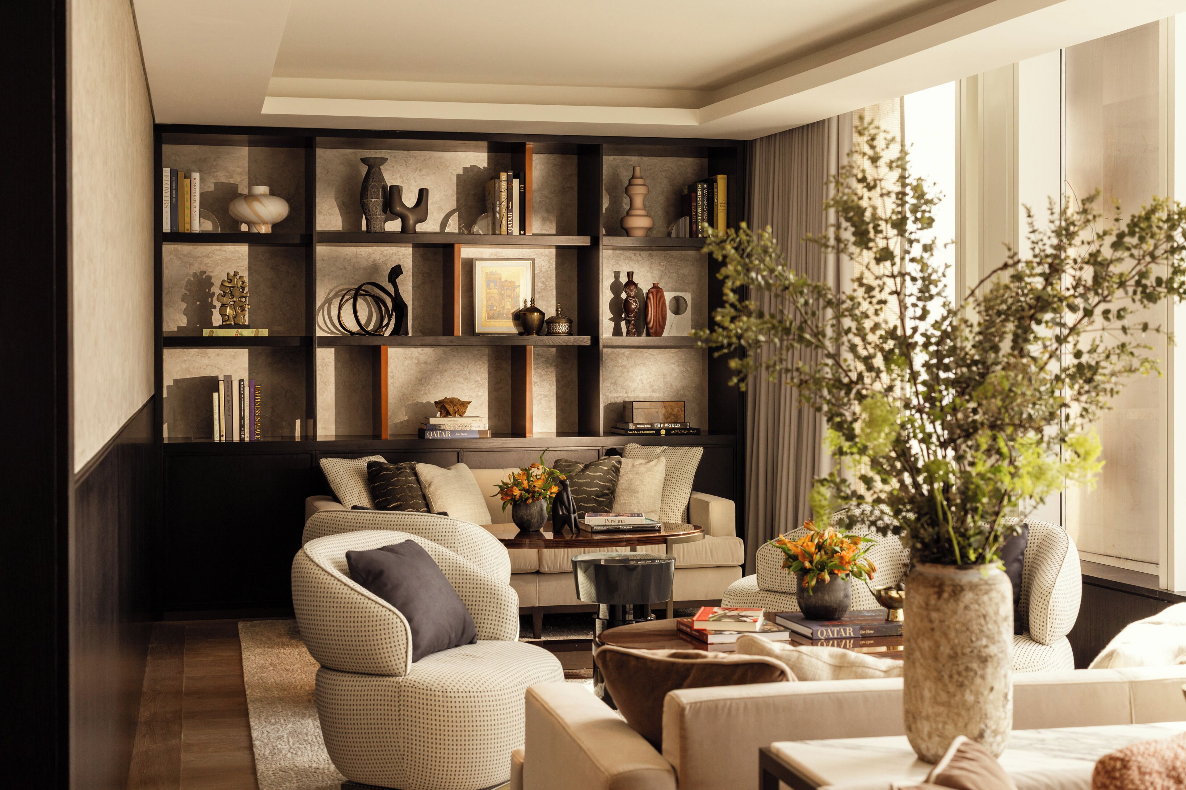 Un salón moderno con una estantería de madera oscura, un sofá de color beige y un amplio ventanal con luz natural.