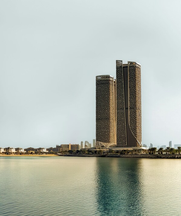 A modern building with a unique facade, situated by a body of water. - Rosewood Doha (Doha)
