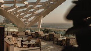 A rooftop terrace with wicker furniture, a glass balustrade, and a view of the ocean. - Rosewood Doha (Doha)