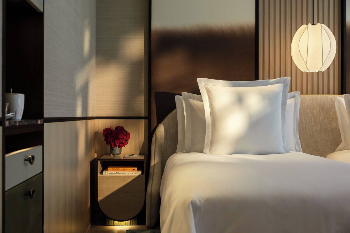 A modern hotel room with a neatly made bed, a bedside table with a vase of flowers, and a wall-mounted lamp.