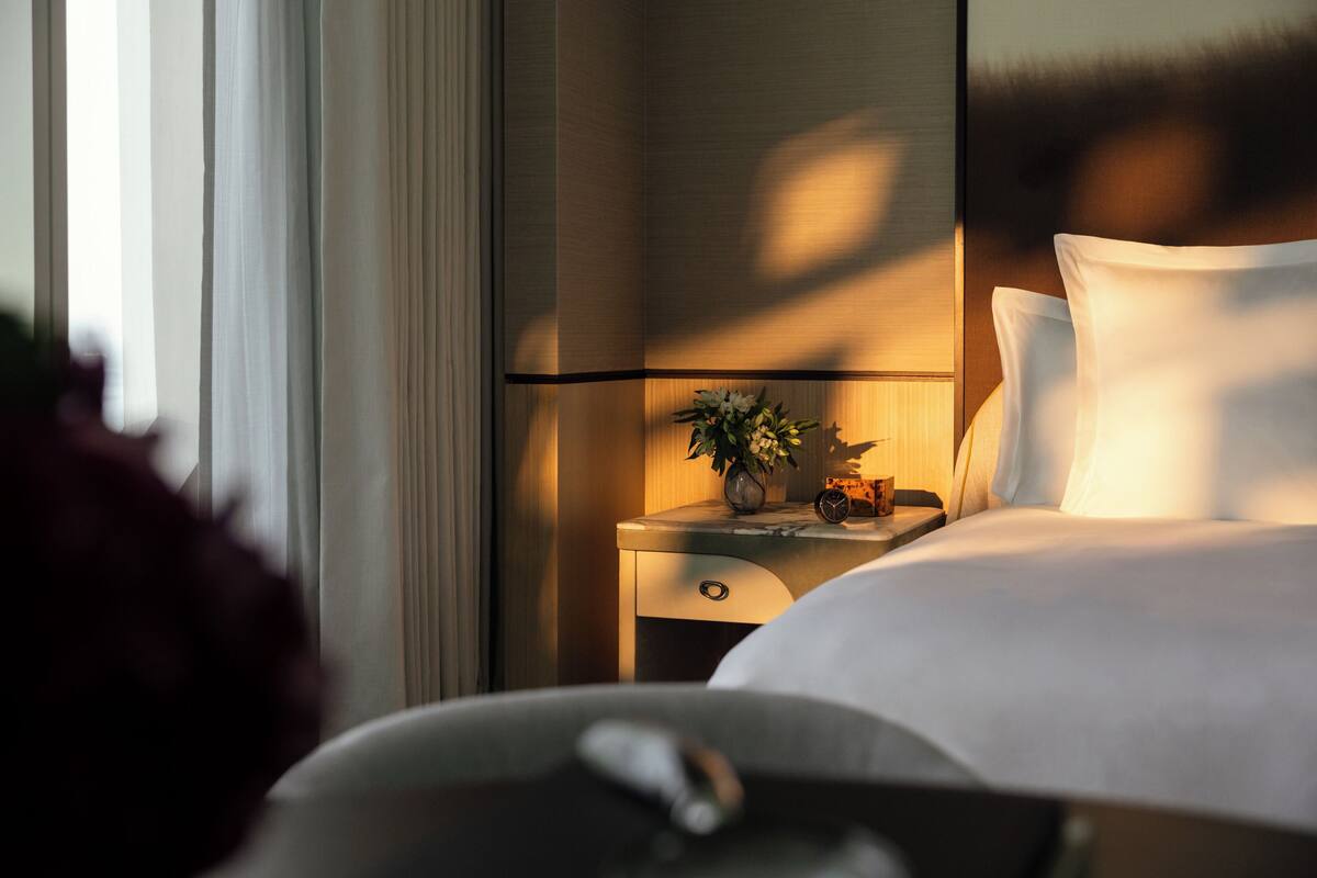 A hotel room with a neatly made bed, a bedside table with a lamp, a clock, and a vase with flowers.