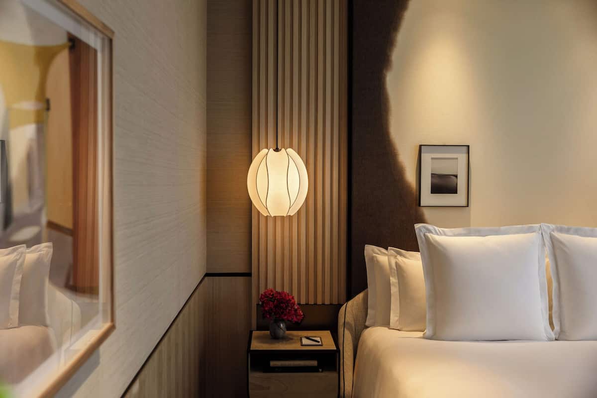A hotel room with a bed, a nightstand with a lamp, a framed picture on the wall, and a window with blinds.