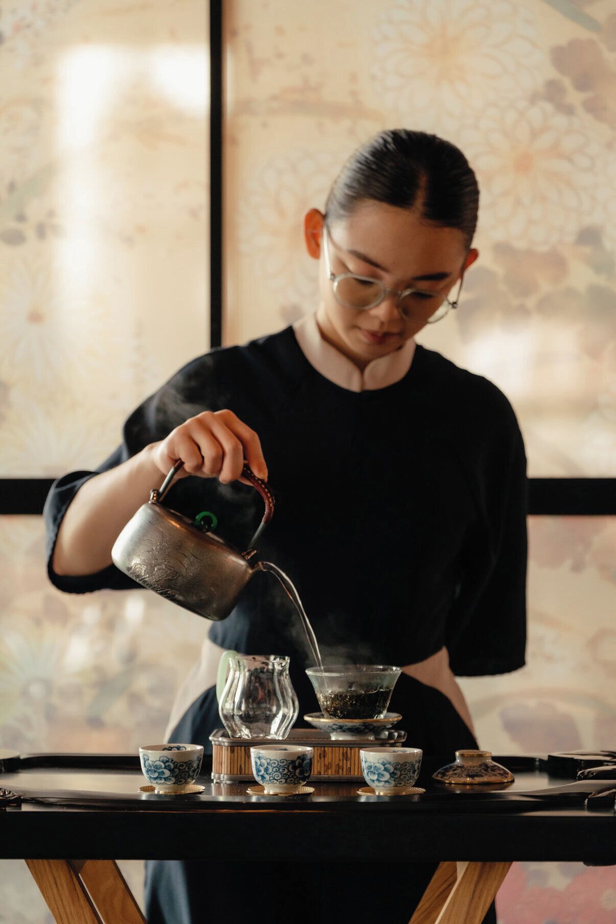 A person pouring tea into a cup on a table with multiple tea sets.