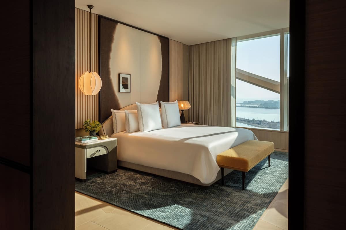A modern hotel room with a large bed, bedside lamps, a view of a body of water, and a cityscape.