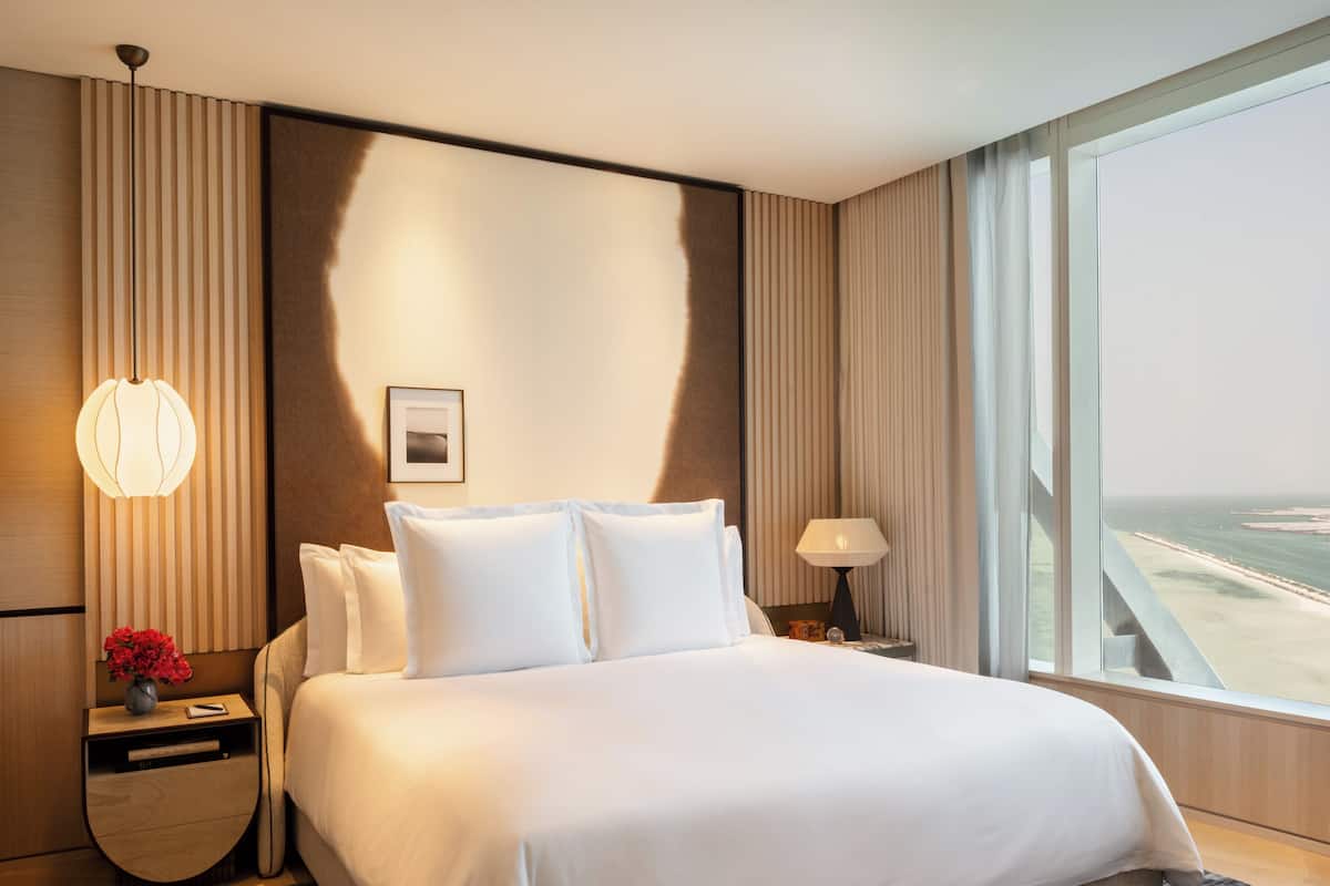 A hotel room with a large bed, a nightstand with a lamp, a vase with flowers, and a view of the beach through the window.