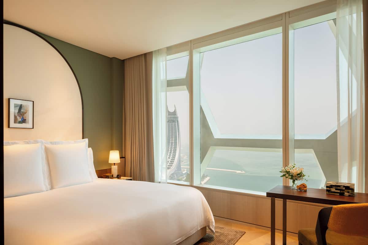A hotel room with a large bed, a desk with a chair, a view of a cityscape through large windows, and a framed picture on the wall.