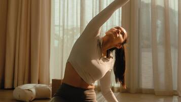 A person in a yoga pose on a mat in a room with large windows and curtains.