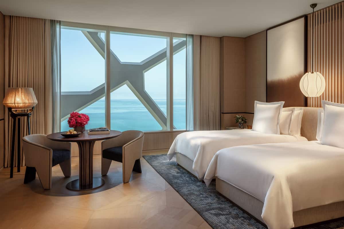 A hotel room with a large window offering a view of the sea, a bed, a desk with a chair, and a lamp.