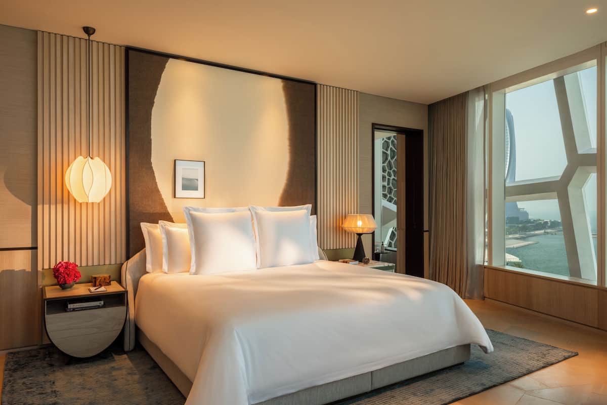 A modern hotel room with a large bed, a nightstand with a lamp, and a view of a cityscape through the window.