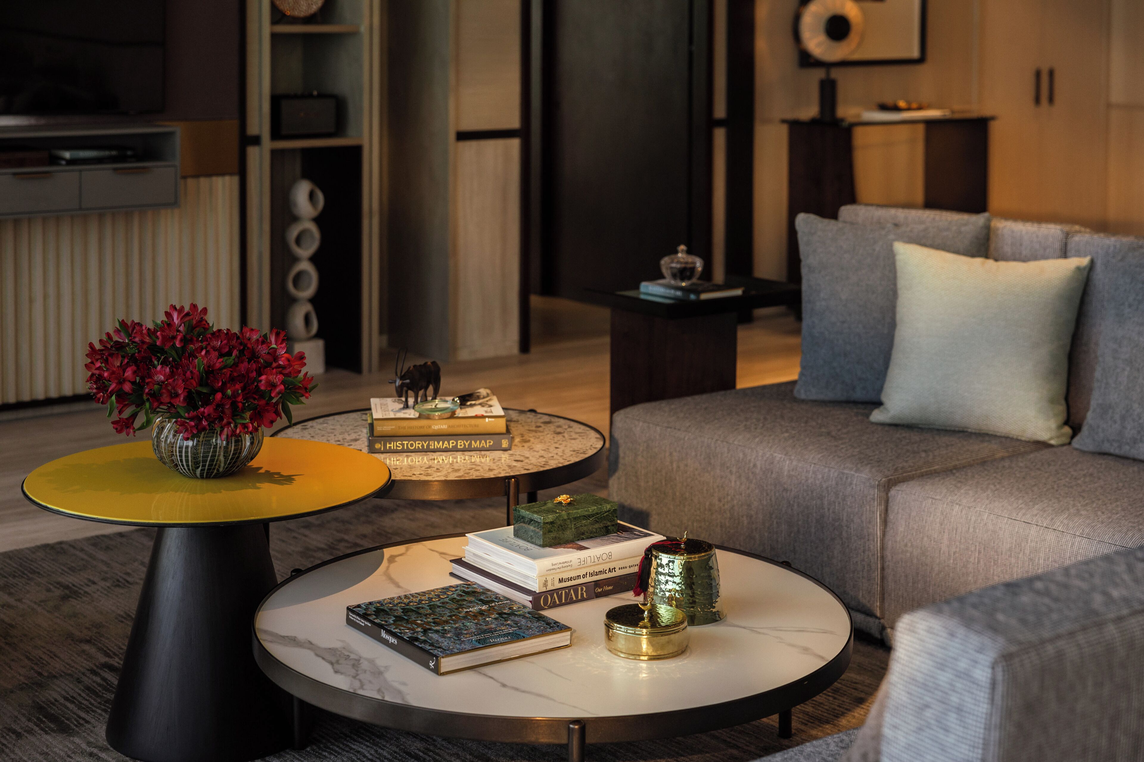 Una sala moderna con un sofá gris, una mesita auxiliar amarilla y negra, y una mesa de centro redonda con libros y objetos decorativos.