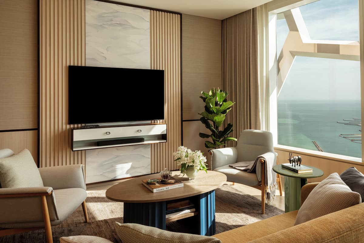 A modern living room with a flat-screen TV, comfortable seating, a coffee table, and a large window with a view of the sea.