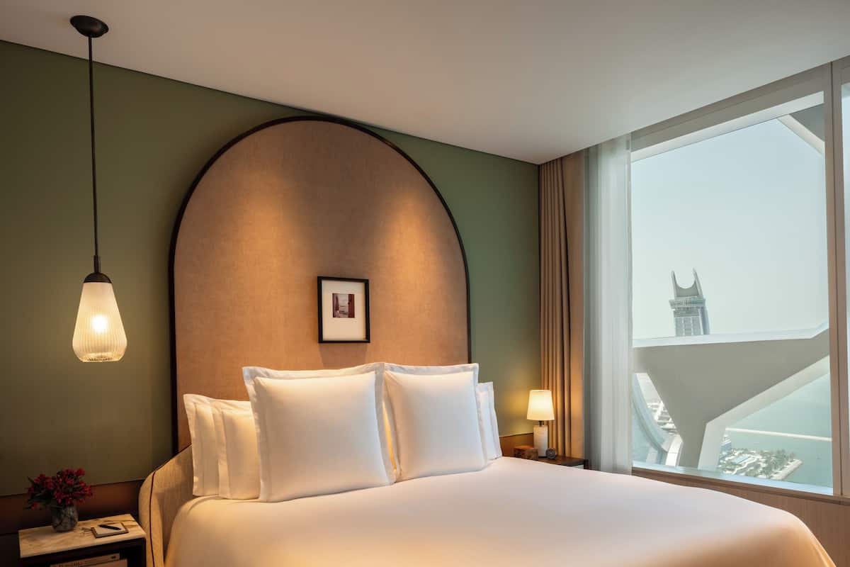 A hotel room with a large bed, a nightstand, a lamp, a framed picture on the wall, and a window with a view of a building.