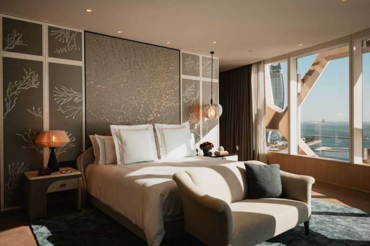 A modern hotel room with a large bed, a sofa, a nightstand, and a view of a cityscape.