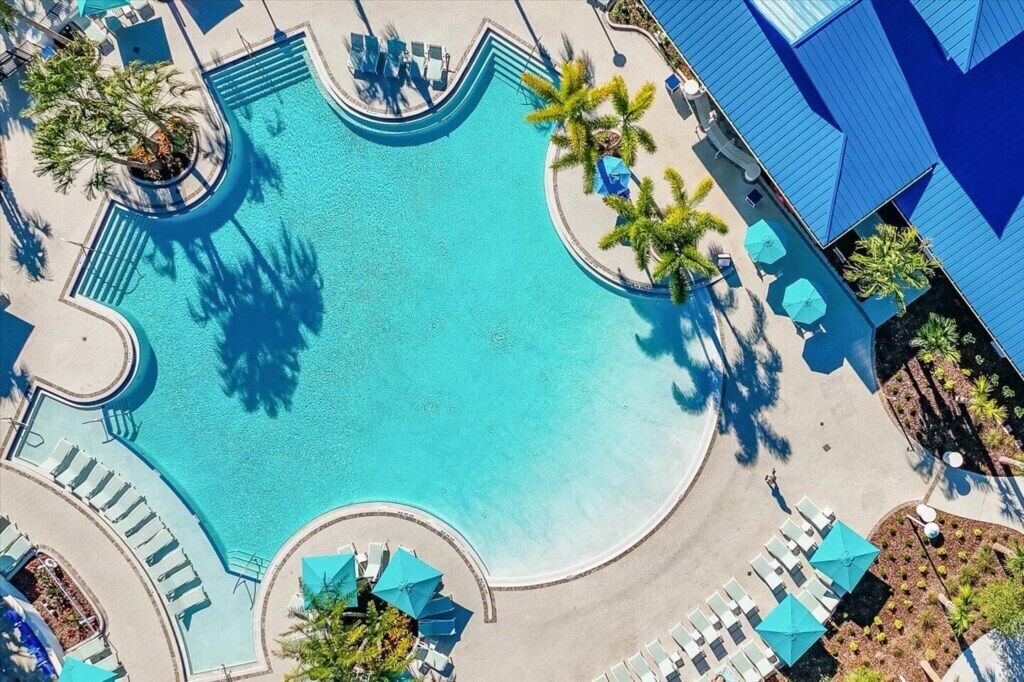 An aerial view of a swimming pool with sun loungers and umbrellas, surrounded by palm trees and a building with a blue roof.