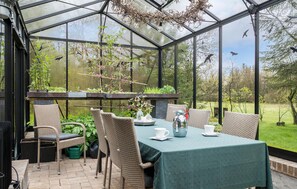 A conservatory with a table set for two, surrounded by wicker chairs, a hanging light fixture, and a view of the garden.