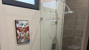 A bathroom with a toilet, a glass shower enclosure, a painting on the wall, and a window above the toilet. - Beautiful Condo with view of the city, mountain & partial Oceanview.  (Playa de oro Zona Hotelera. Nte.c p)
