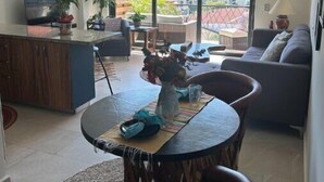 A modern living room with a dining area, a sofa, a coffee table, and a TV. There is a large window offering a view of the outdoors. - Beautiful Condo with view of the city, mountain & partial Oceanview.  (Playa de oro Zona Hotelera. Nte.c p)