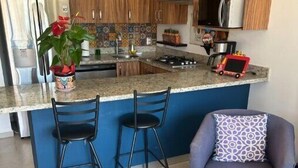 A kitchen with wooden cabinets, a granite countertop, a gas stove, and a red toy truck. There are two black bar stools and a purple armchair with a patterned pillow. - Beautiful Condo with view of the city, mountain & partial Oceanview.  (Playa de oro Zona Hotelera. Nte.c p)