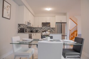 Una cocina moderna con armarios blancos, salpicadero de azulejos y electrodomésticos de acero inoxidable. Hay una mesa de comedor de vidrio con sillas blancas.