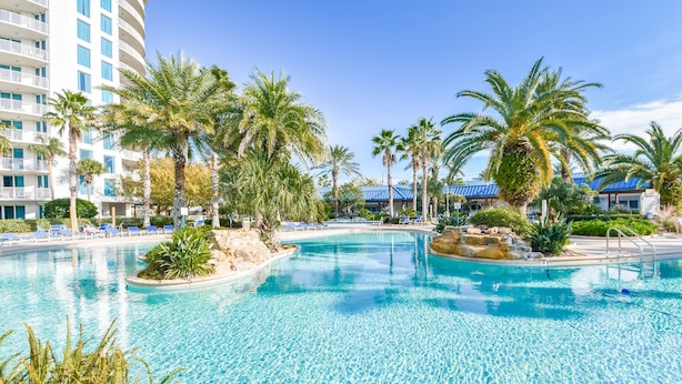 A large outdoor swimming pool surrounded by palm trees and lounge chairs, with a hotel building in the background. - Salted Tranquility at The Palms Of Destin (Destin)