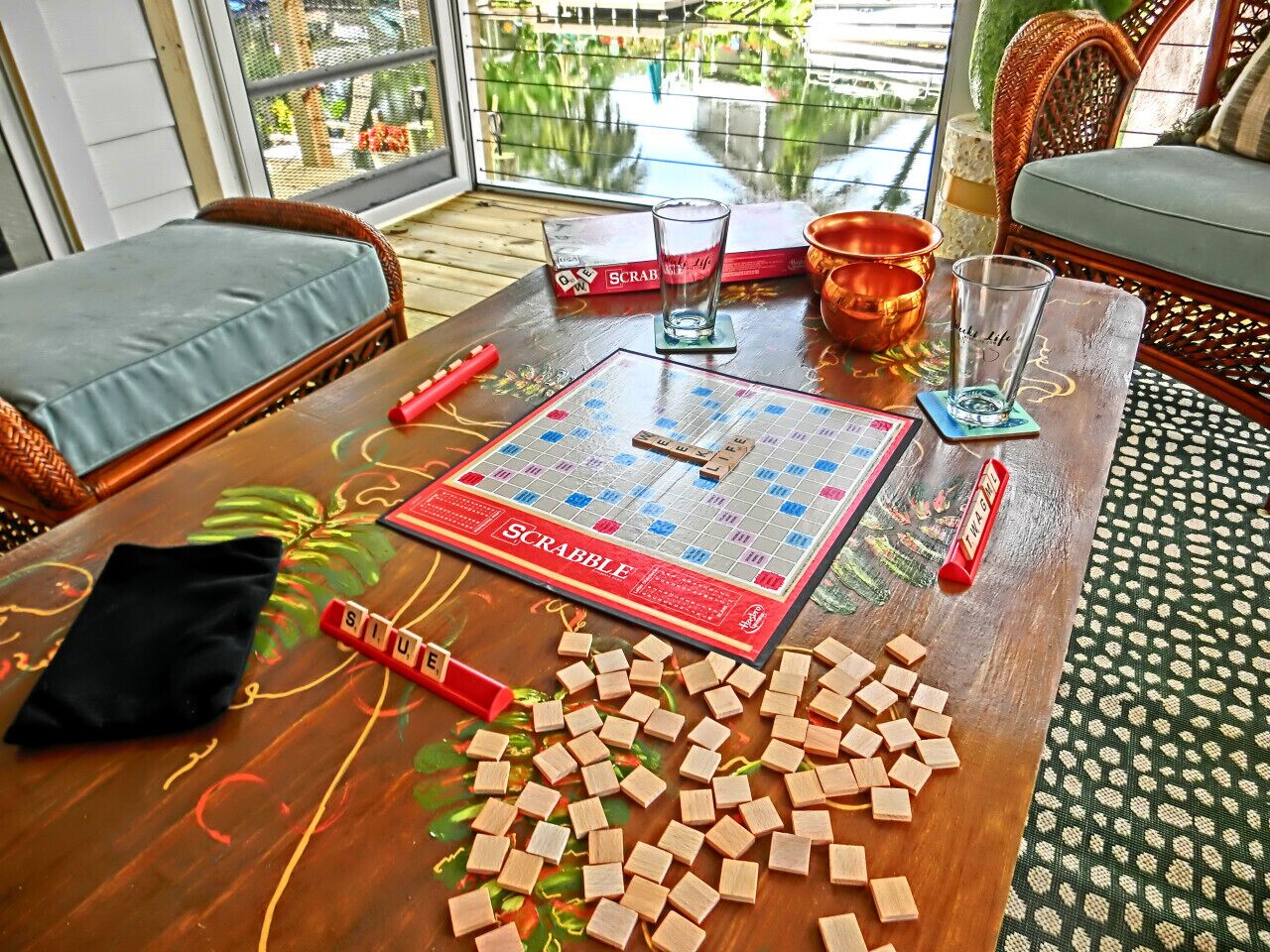 A Scrabble game on a wooden table with scattered tiles, a Scrabble box, and two glasses.