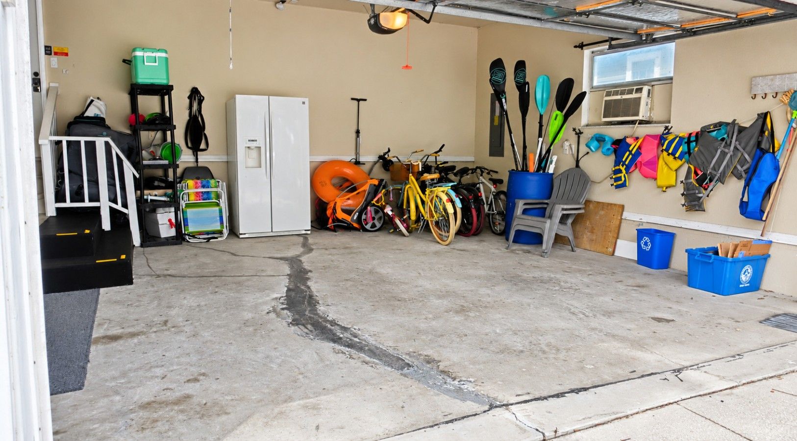 A garage with a concrete floor, a white refrigerator, a bicycle, kayaks, and various storage bins.