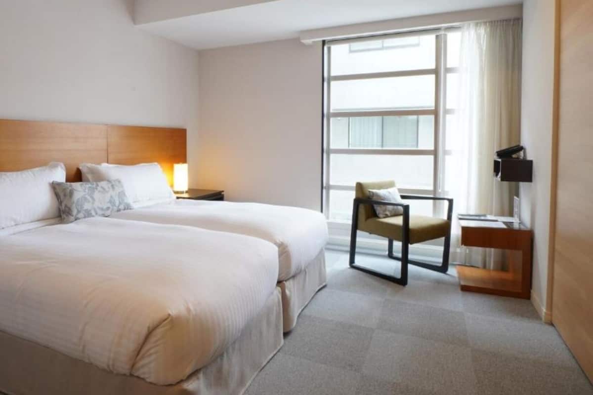 A hotel room with two beds, a chair, a desk, and a large window.