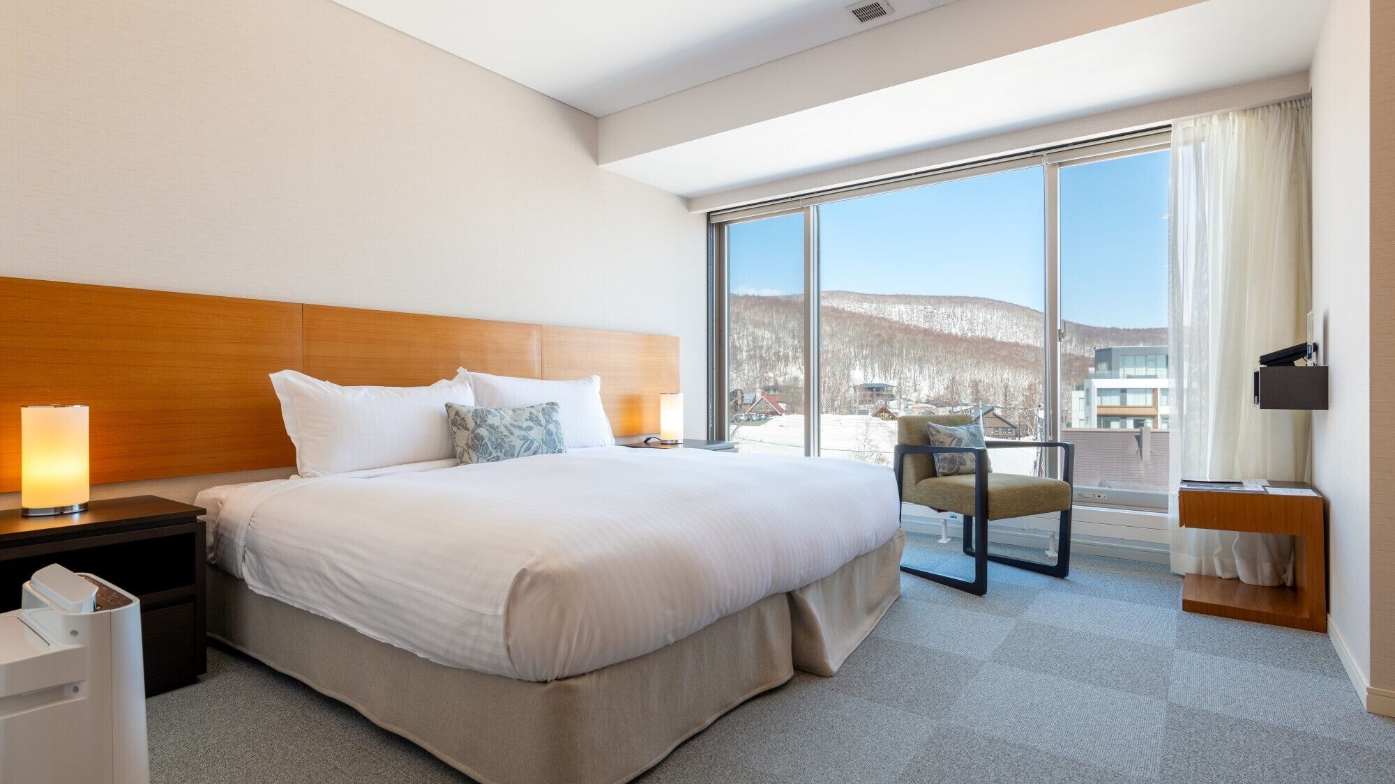 A hotel room with a large bed, a nightstand, a chair, a desk with a lamp, and a large window with a view of a snowy landscape.