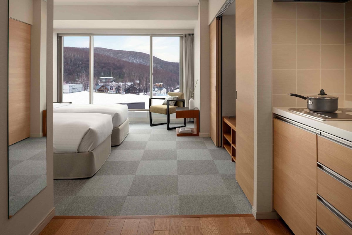 Una habitación de hotel con un ventanal grande, una cama, una silla, una mesita y una kitchenette.