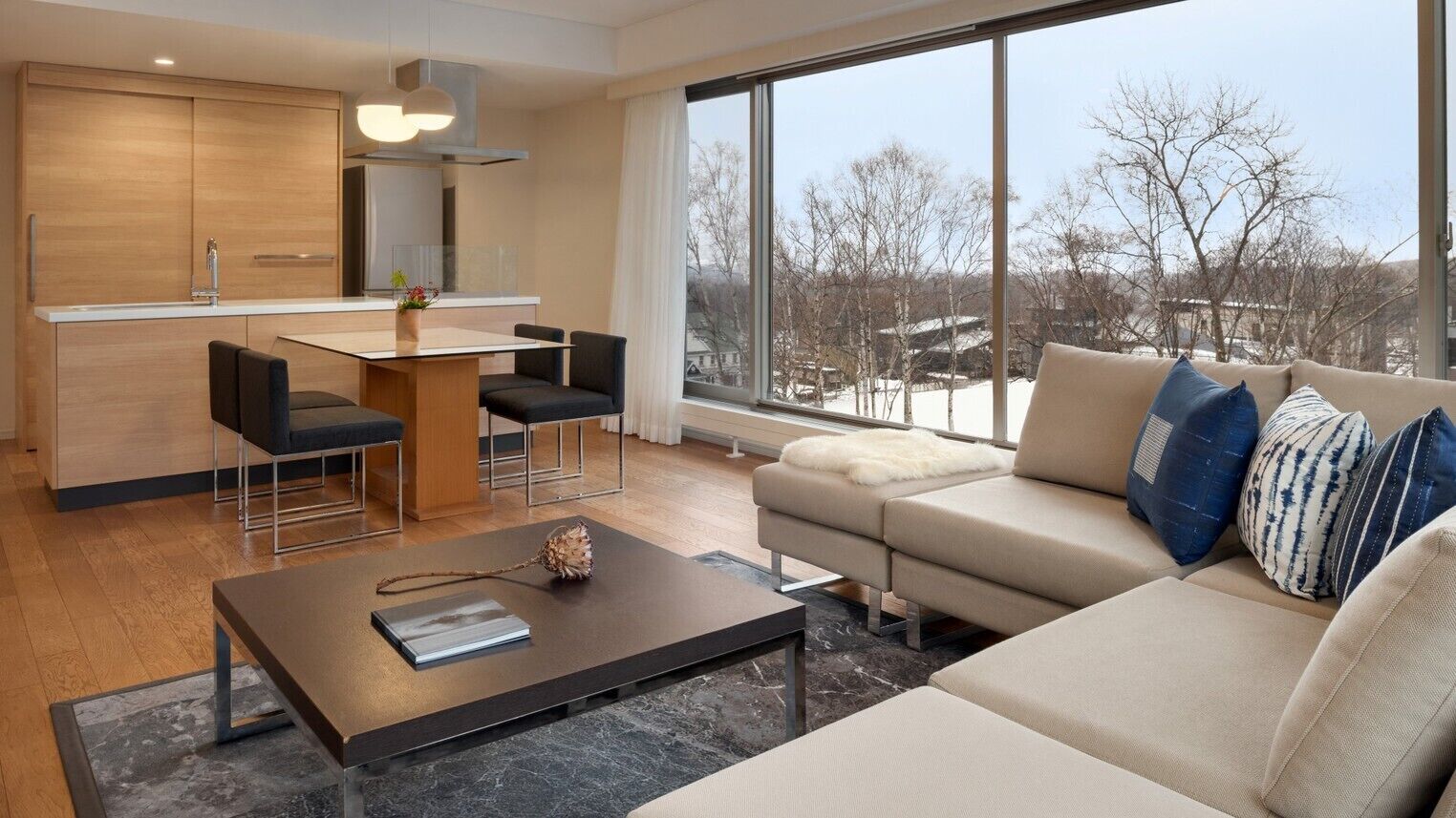 A modern living space with a kitchen, dining area, and a large window offering a view of trees and buildings.