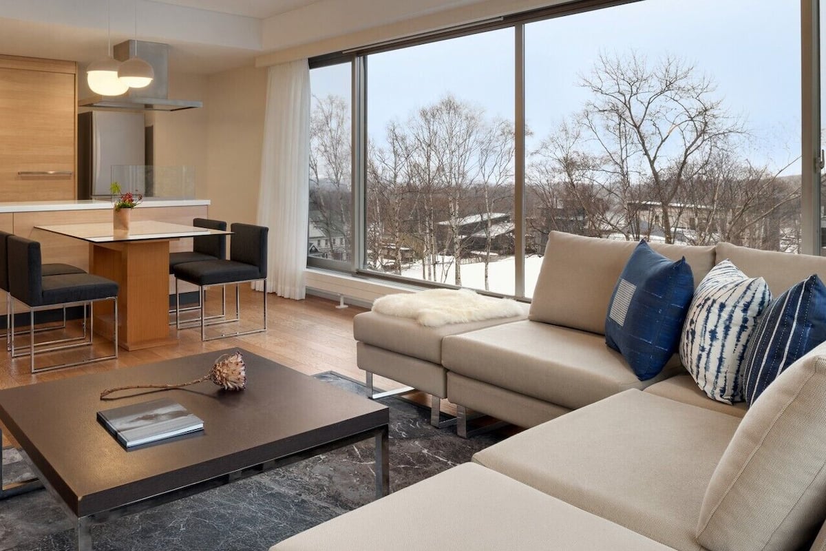 Un espacio habitable moderno con cocina, comedor y un gran ventanal que ofrece vistas a árboles y edificios.
