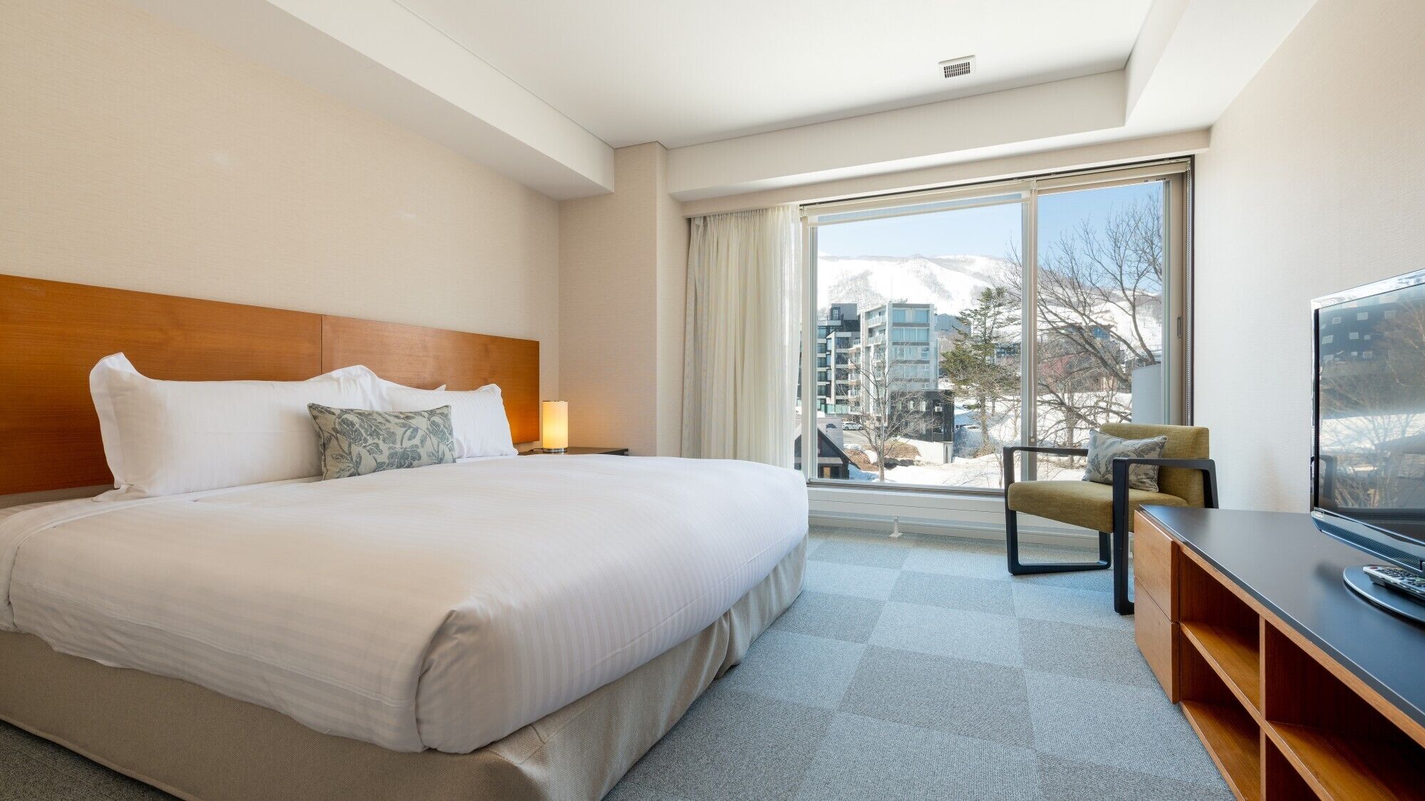 A hotel room with a large bed, a TV, a desk, and a window with a view of buildings and snow.