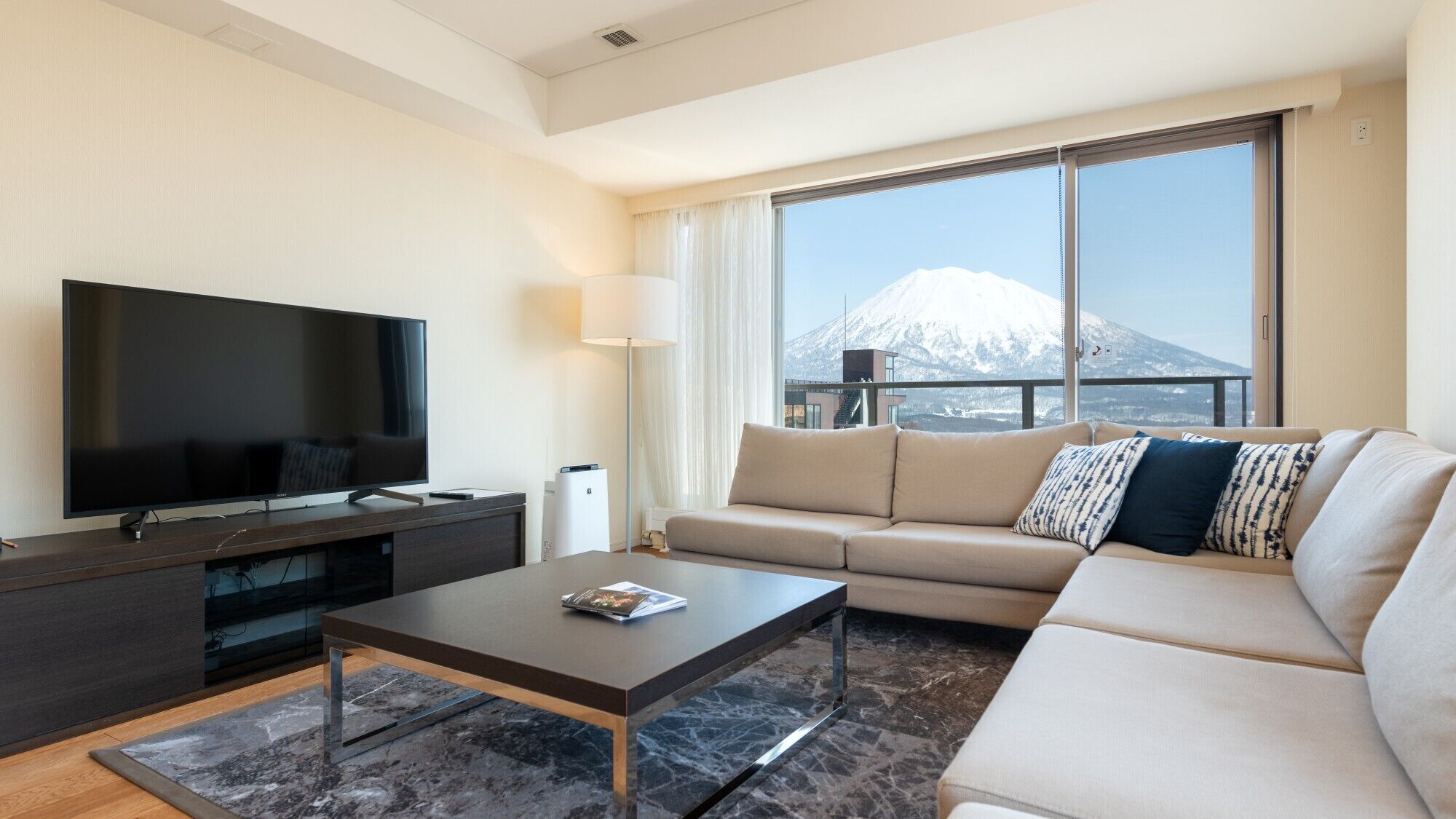 A modern living room with a flat-screen TV, a sofa, a coffee table, and a large window with a mountain view.
