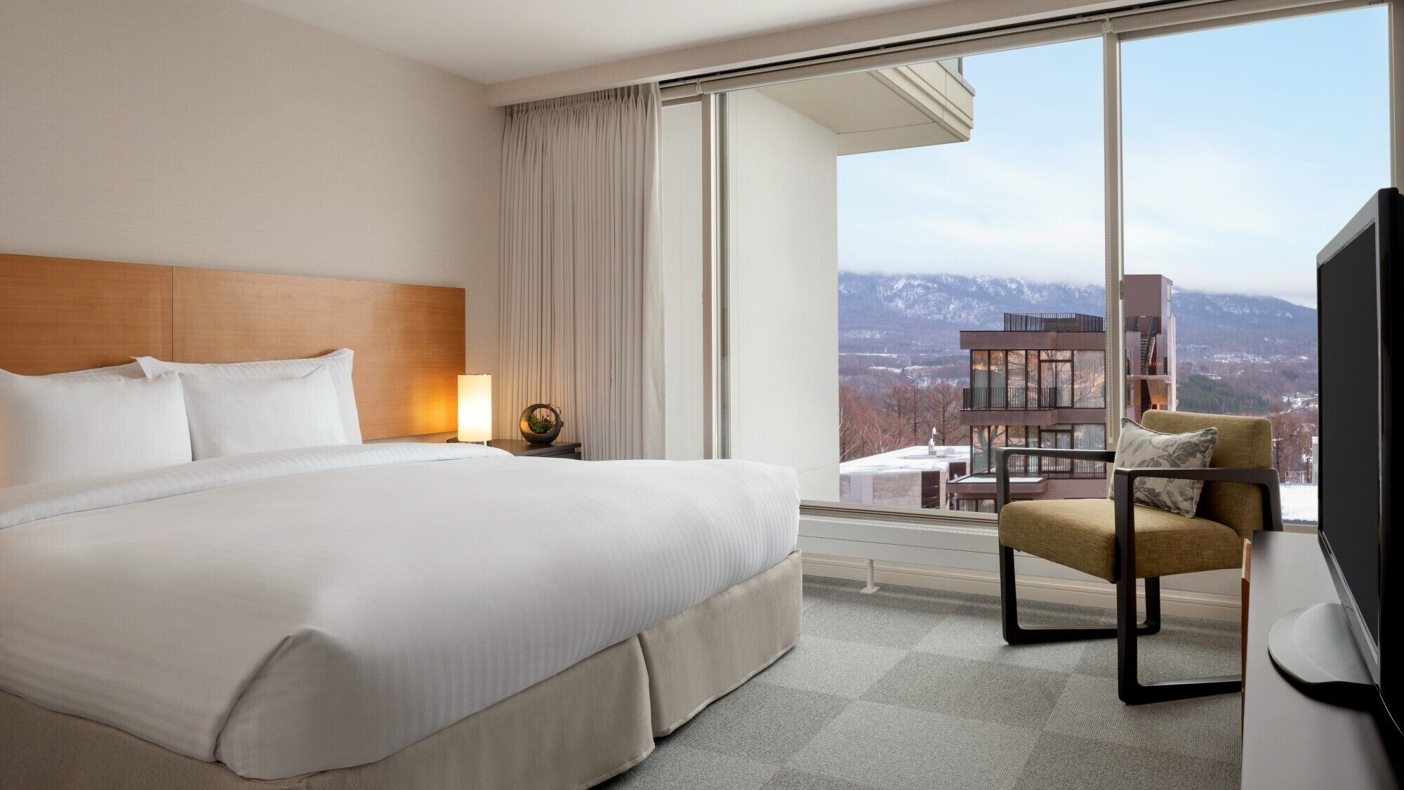 A hotel room with a large bed, a chair, a TV, and a view of mountains.
