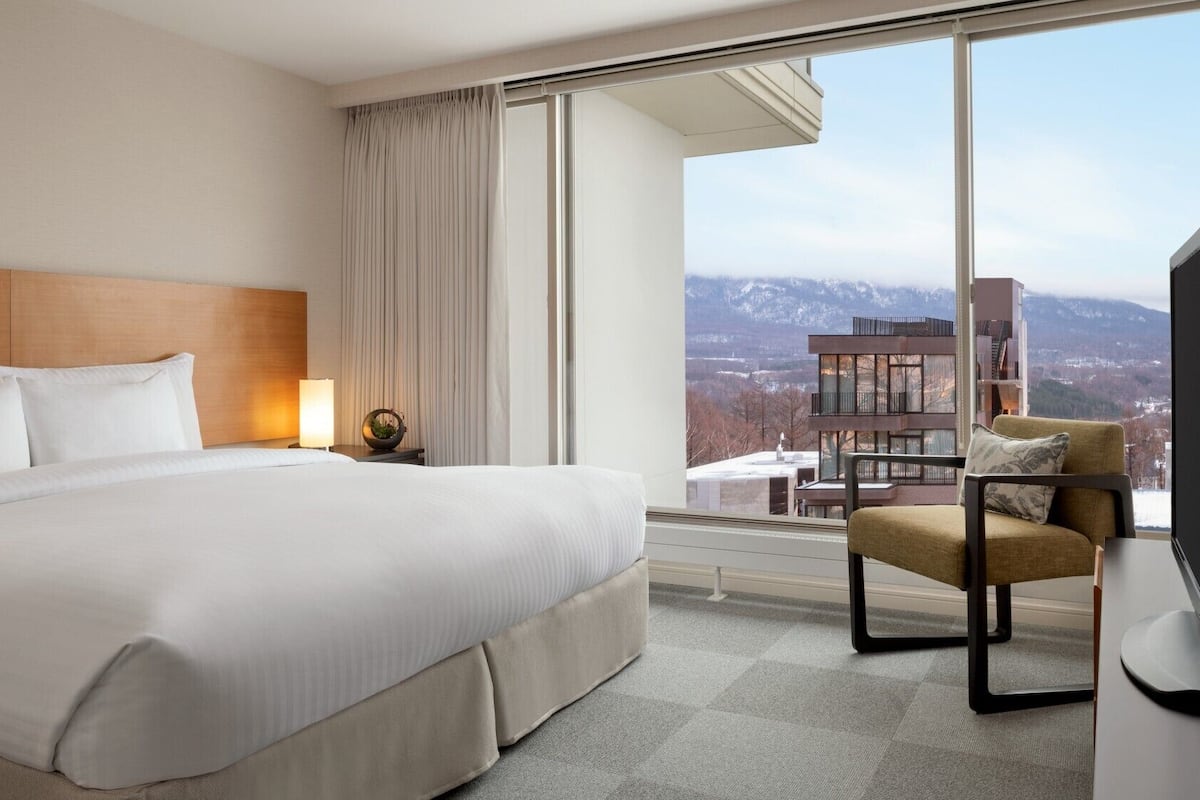 Habitación de hotel con una cama grande, una silla, un televisor y vistas a las montañas.