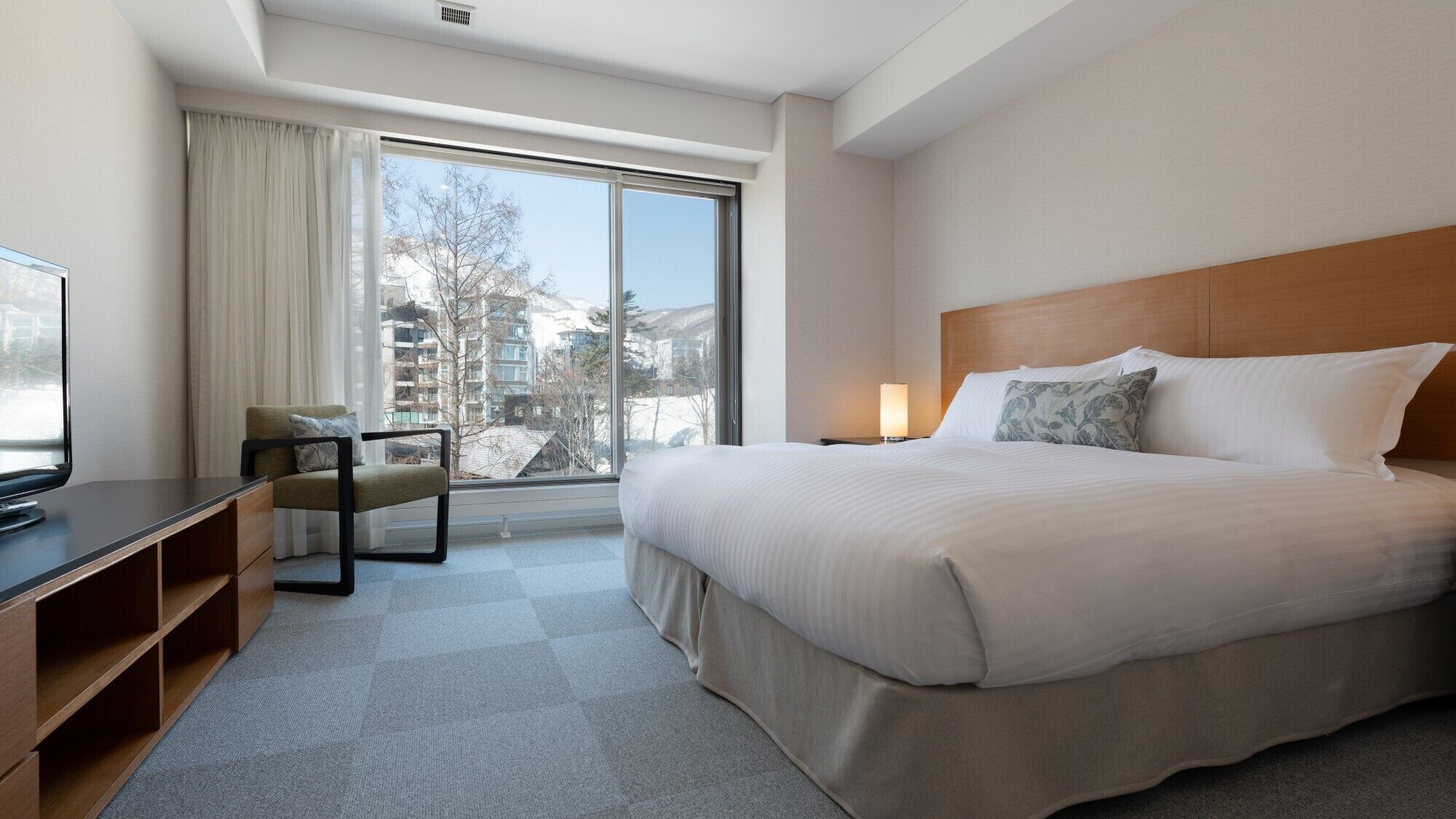 A hotel room with a bed, a desk with a TV, a chair, a nightstand, and a window with a view of buildings and trees.