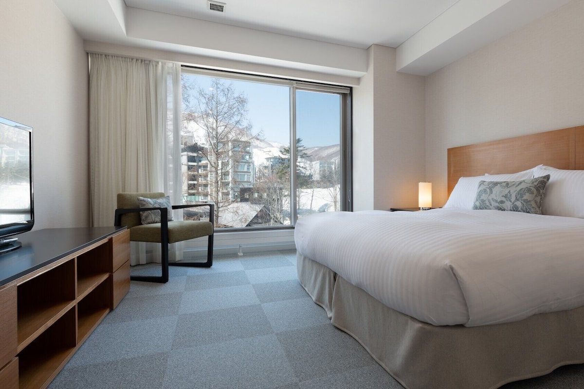 Una habitación de hotel con una cama, un escritorio con televisor, una silla, una mesita de noche y una ventana con vistas a edificios y árboles.