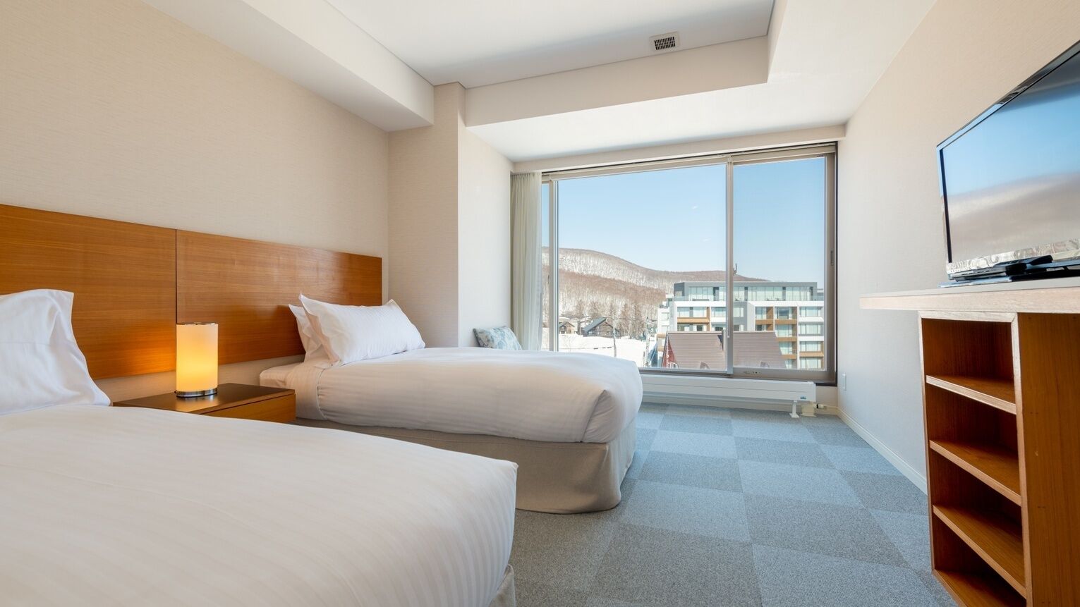 A hotel room with two beds, a TV, a large window, and a view of buildings.
