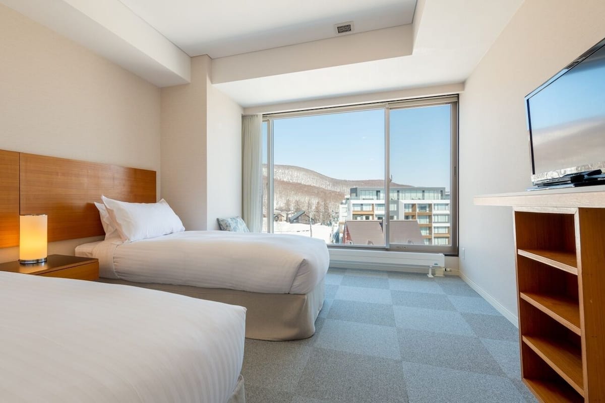 Una habitación de hotel con dos camas, un televisor, un ventanal grande y vistas a edificios.