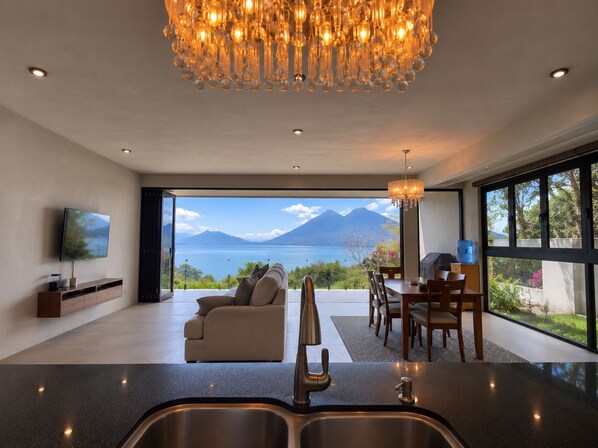 A modern living room with a large window, a sofa, a dining area, and a kitchen with a sink.