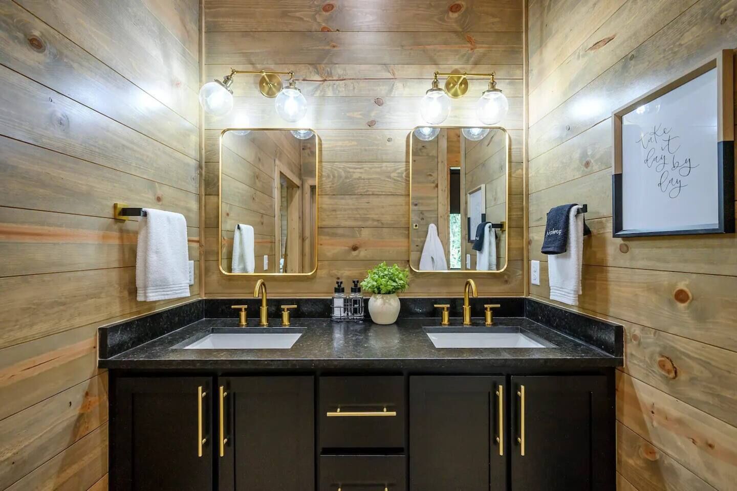 A wooden-paneled bathroom with two sinks, gold fixtures, and a mirror with lights.