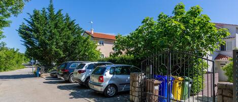 A row of parked cars along a paved road with a metal fence and trash bins nearby.