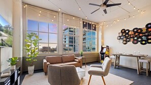 Un salon moderne comprenant un canapé, des fauteuils, une table basse et une table à manger avec des chaises. De grandes fenêtres, un ventilateur de plafond et des guirlandes lumineuses complètent l’ensemble.